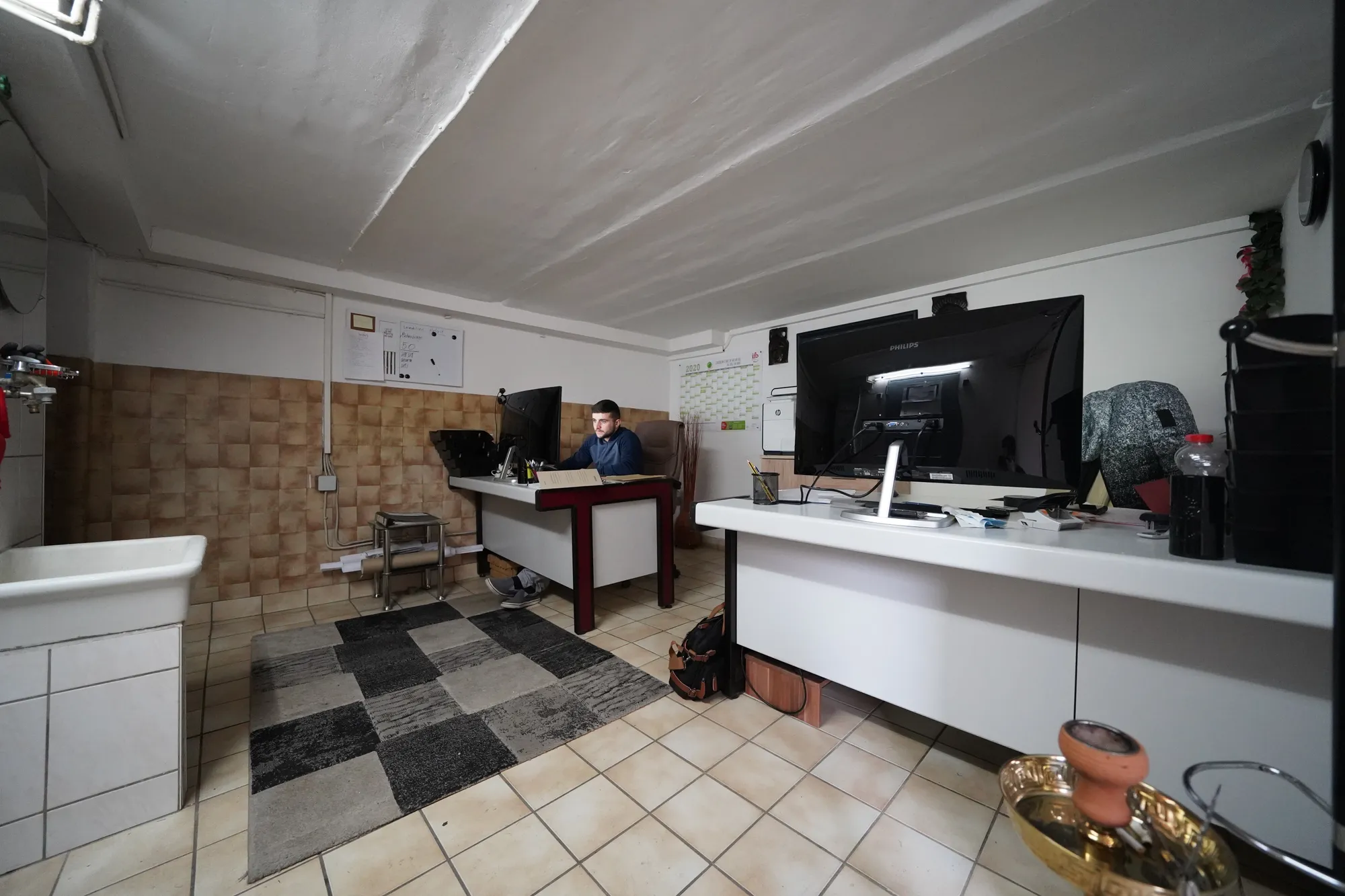 Der erste Arbeitsplatz im Keller: ein Schreibtisch, zwei Bildschirme, Fliesenboden und ein Waschbecken